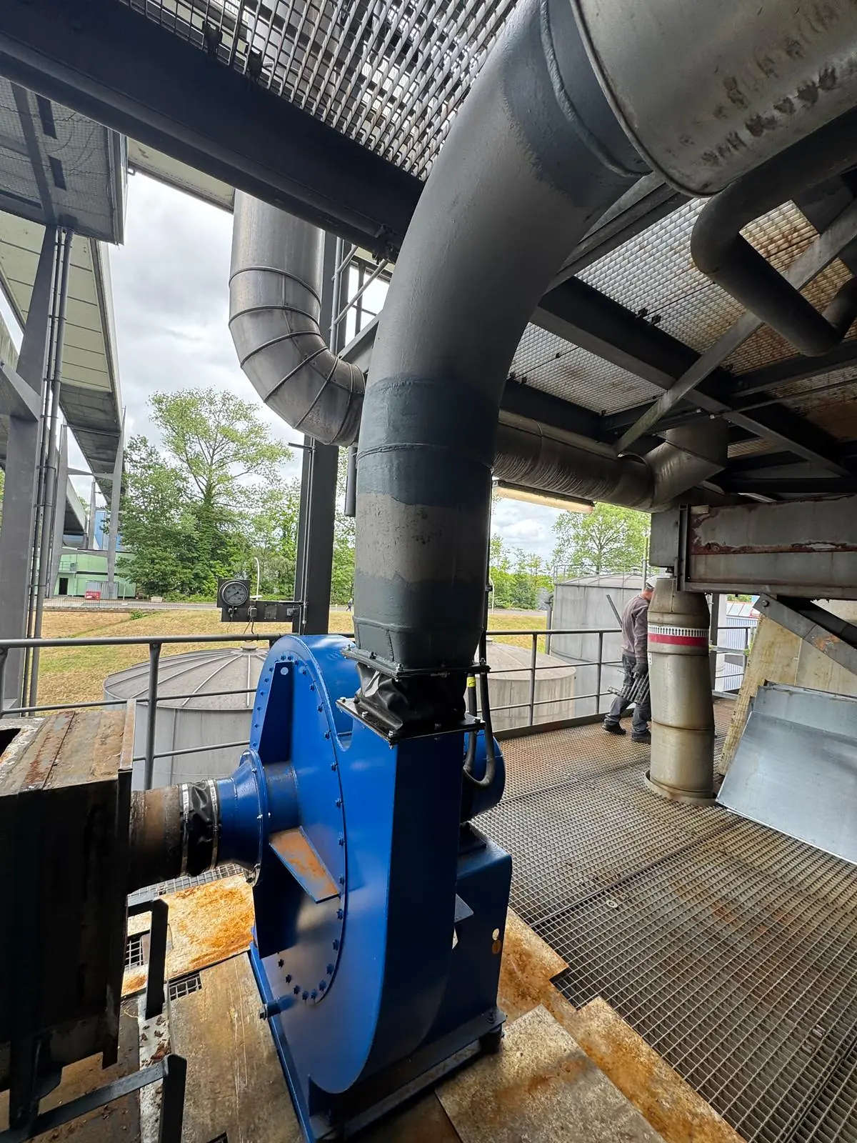 Nuovo ventilatore centrifugo blu installato su un basamento in acciaio su una piattaforma a grigliato all’aperto, con la mandata collegata tramite un giunto di dilatazione al camino di estrazione esistente verniciato