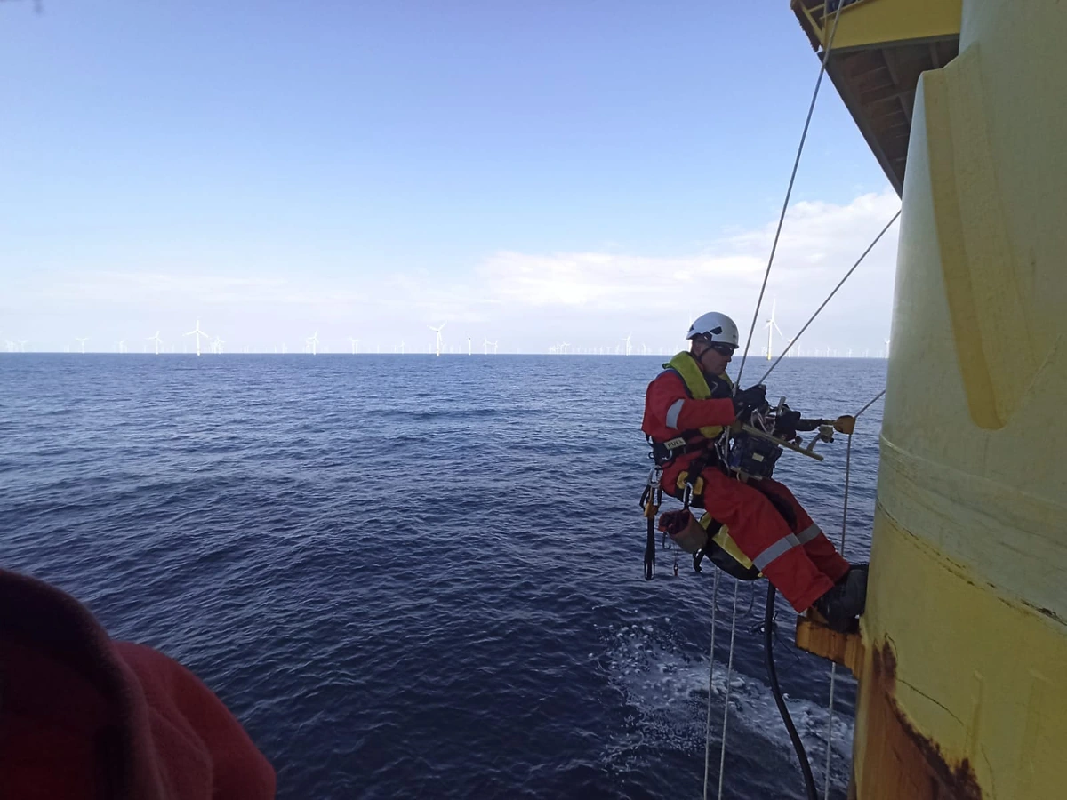 Technicien cordiste suspendu dans son harnais sur la face extérieure d’une plateforme offshore, éoliennes à l’horizon