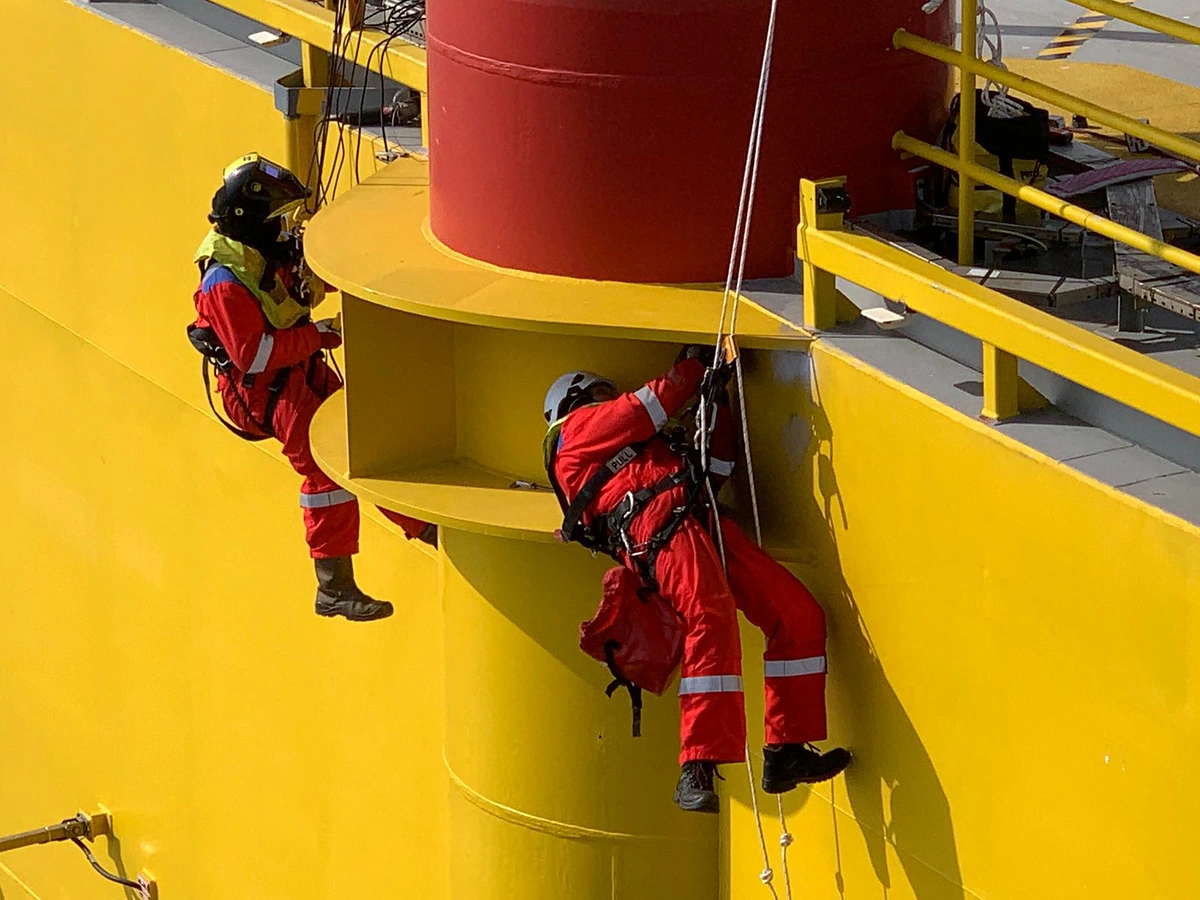 Deux techniciens cordistes suspendus sur cordes jumelées à une structure jaune d’une plateforme offshore, l’un portant un casque de soudage, les câbles descendus depuis le pont au-dessus