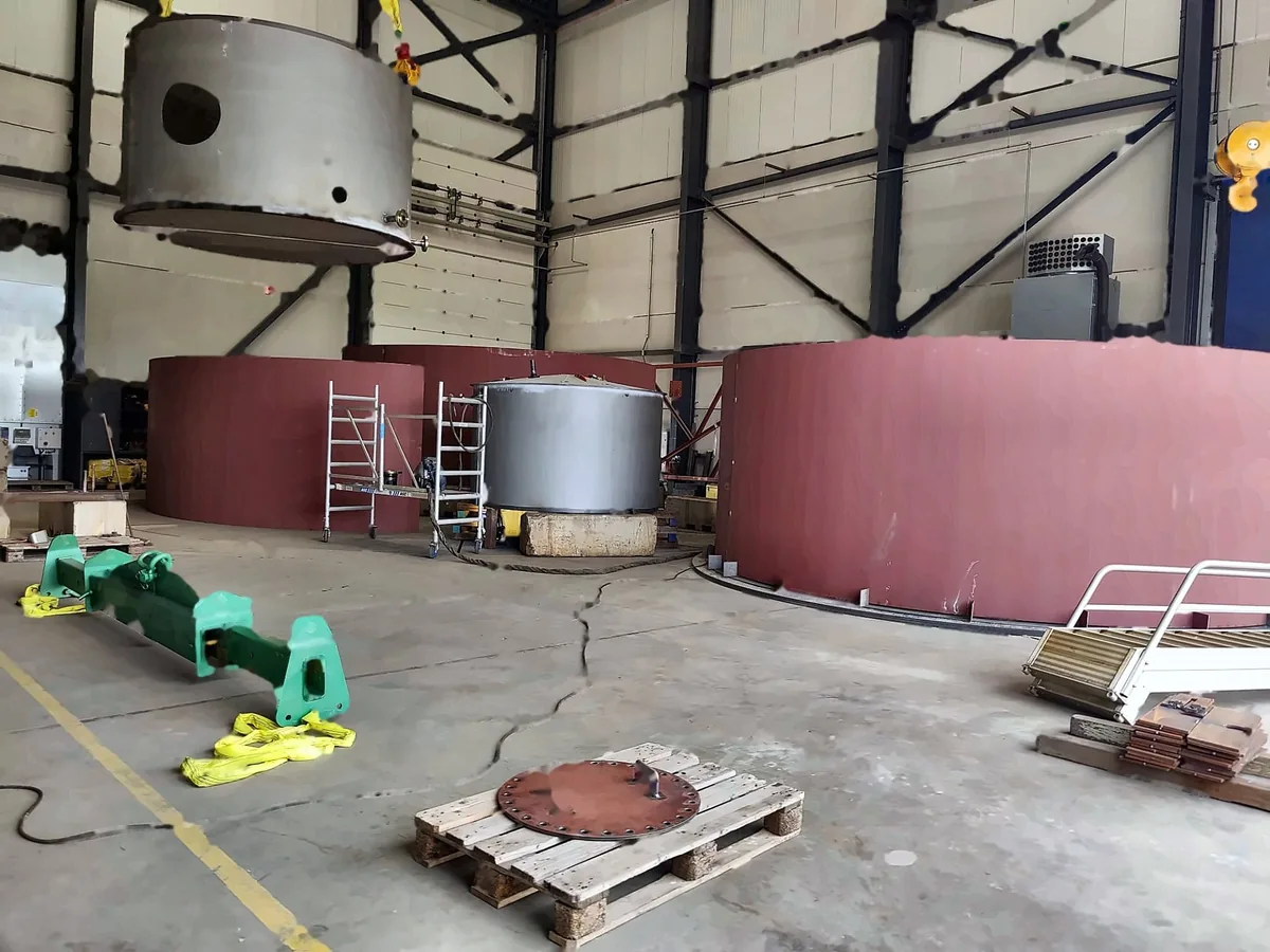 Fertigungshalle mit Tankmänteln, einem Behälter auf Holzunterbau und einer Traverse mit Anschlagmitteln am Boden